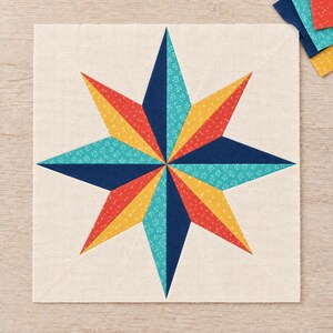 May include: A square fabric quilt block featuring a central eight-pointed star design. The star is composed of alternating segments in navy blue, teal with a white floral pattern, yellow with white dots, and red with white dots on a cream background.