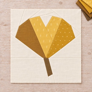 May include: A square fabric quilt block featuring a stylized flower design in shades of mustard yellow, gold, and brown. The flower petals are composed of solid, dotted, and sprig-patterned fabric pieces set against a cream-colored background.