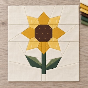 May include: A square fabric quilt block featuring a pieced sunflower design. The flower has a dark brown center with small dots, yellow petals with subtle white sprig patterns, and a green stem with two leaves, all set against a cream background.