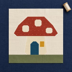 May include: A square fabric quilt block featuring a pieced mushroom house design. The mushroom cap is red with white polka dots and white octagonal spots. The stem is cream with a blue door and a yellow window, set against a green ground.