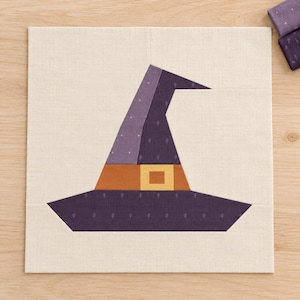 May include: A square fabric quilt block featuring a pieced witch hat design. The hat is constructed from dark purple and light purple fabric with a small white leaf pattern, an orange band, and a yellow square buckle, all on a cream background.