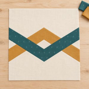 Könnte beinhalten: Ein quadratischer Stoff-Quiltblock mit cremefarbenem Hintergrund und einem geometrischen Zickzackmuster. Das Design besteht aus zwei sich kreuzenden Bändern: eines in Senfgelb und eines in Petrolblau mit einem kleinen weißen Blattmuster.