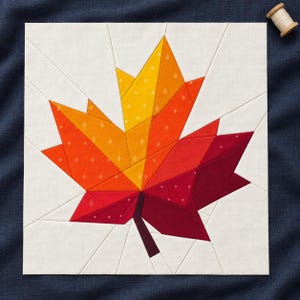 May include: A square fabric quilt block featuring a paper-pieced maple leaf design. The leaf transitions from yellow to orange to deep red, set against a white background. The fabric pieces have small white patterns and a dark brown stem.