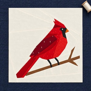 May include: A square fabric quilt block featuring a geometric paper-pieced cardinal bird in shades of bright red and dark maroon. The bird has a black face mask, an orange beak, and sits on a brown branch against a solid off-white background.
