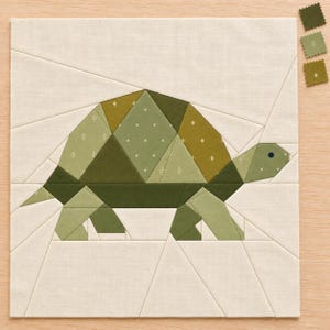 Könnte beinhalten: Ein quadratischer Stoff-Quiltblock mit einem geometrischen Schildkrötenmotiv, das aus verschiedenen grünen Baumwollstoffen zusammengesetzt ist. Die Schildkröte ist auf einem cremefarbenen Hintergrund mit sichtbaren Nähten und drei Stoffmustern.