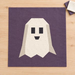 May include: A square fabric quilt block featuring a stylised white ghost shape centred on a solid dark purple background. The ghost has two black square eyes, a small black triangular nose, and light grey side panels with a subtle dot pattern.