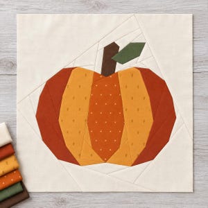 May include: A square fabric quilt block featuring a paper-pieced pumpkin design. The pumpkin is constructed from segments of orange, rust, and yellow fabric with small white polka dot and leaf patterns, a dark brown stem, and a green leaf.
