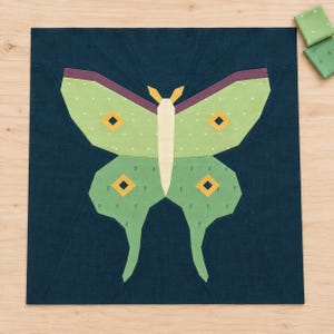 May include: A square fabric quilt block featuring a central moth design in shades of light green, cream, and dark blue. The moth has four wings with yellow diamond-shaped accents, a cream-colored body, and dark purple wing tips on a dark blue background.