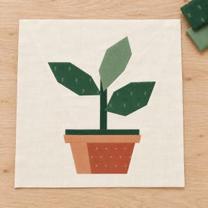 May include: A square fabric quilt block featuring a potted plant design. The plant has a dark green stem and three geometric leaves in varying shades of green with small white sprig patterns. The pot is terracotta orange with white dots.
