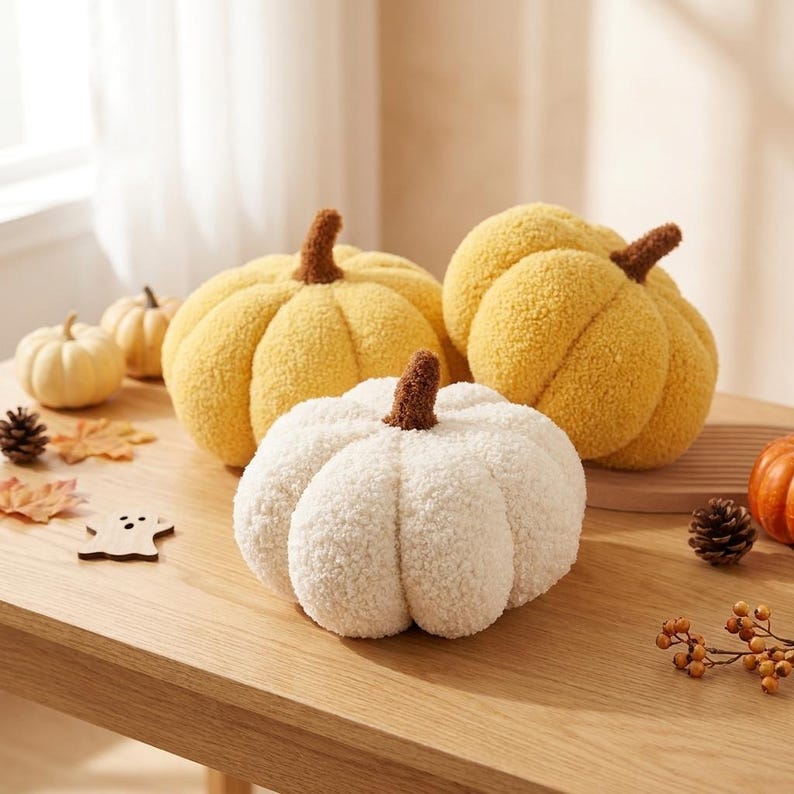 May include: Three plush pumpkin-shaped pillows with a textured, sherpa-like fabric. One pillow is white, one is light yellow, and one is a deeper golden yellow. Each pumpkin features a small, brown, fuzzy stem at the top center of the rounded body.