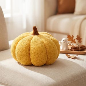 May include: A round, mustard yellow pumpkin-shaped pillow with a textured, fuzzy surface and a small, brown, nubby stem at the top. The pillow features deep, vertical indentations that define its segments, creating a soft, plush appearance.