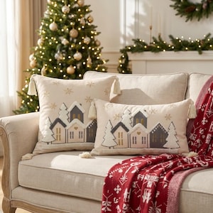 May include: Two cream-coloured throw pillows featuring embroidered winter village scenes with houses, trees, and snowflakes. Each pillow is accented with decorative tassels at the corners and displays a soft, textured design on the front surface.