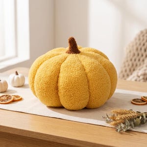 May include: A round, textured yellow pumpkin-shaped pillow with a brown stem. The fabric has a soft, fuzzy, sherpa-like appearance with deep vertical ridges creating a segmented, realistic gourd shape. The pillow is a solid, vibrant mustard yellow.