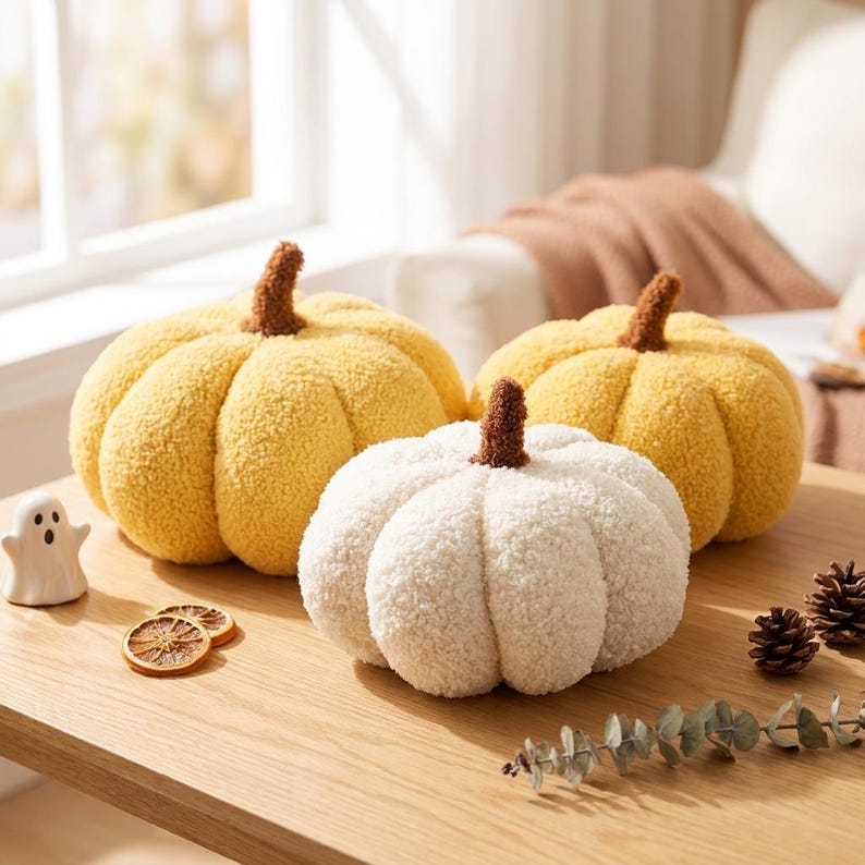 May include: Three plush pumpkin-shaped pillows with textured, fuzzy fabric. Two pumpkins are yellow and one is white, each featuring a brown stem. The set includes a small ceramic ghost figurine, two dried orange slices, and pinecones.