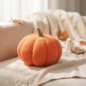 May include: A round, orange pumpkin-shaped pillow with a textured, fuzzy surface and a small, upright stem in a muted brown color. The pumpkin features deep, vertical segments that create a realistic, rounded shape for this decorative item.