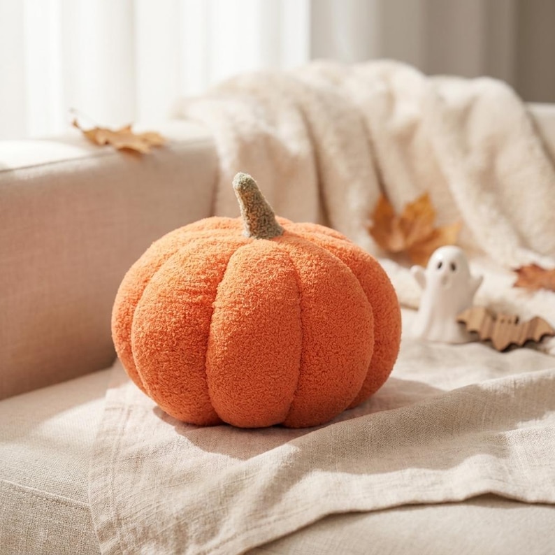 May include: A round, orange pumpkin-shaped pillow with a textured, fuzzy surface and a small, upright stem in a muted brown color. The pumpkin features deep, vertical segments that create a realistic, rounded shape for this decorative item.