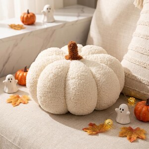 May include: A round, cream-colored, textured sherpa pumpkin pillow with a brown stem. The pumpkin features deep, defined segments and a soft, fuzzy surface. It is surrounded by small white ghost figurines, orange pumpkins, and orange autumn leaves.