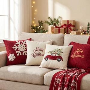 May include: Four square throw cushions arranged on a sofa. One red cushion features white snowflake appliques. A cream cushion displays the red embroidered text Merry Christmas. Another cream cushion shows a red car carrying a Christmas tree. A red cushion features gold embroidered text Merry Christmas.
