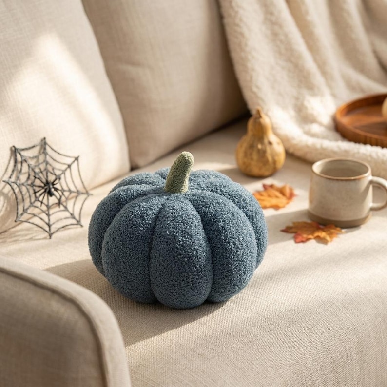 May include: A round, blue, textured pumpkin-shaped pillow with a small, light green stem. The fabric has a soft, fuzzy, boucl&eacute;-like appearance with deep vertical ridges that define the segments of the pumpkin shape, creating a plush, rounded form.