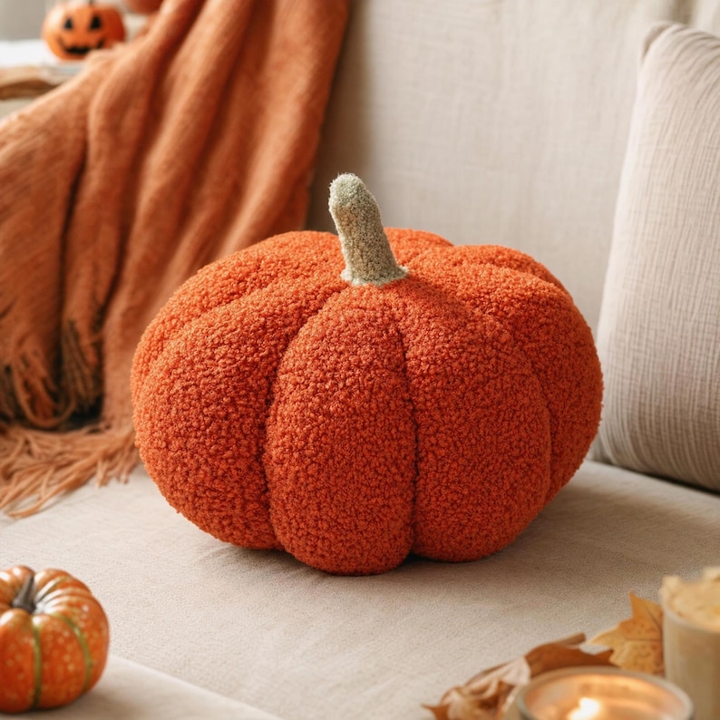 May include: A round, orange pumpkin-shaped pillow made of a textured, fuzzy fabric. The pillow features deep vertical ridges and a short, light green stem at the top center. The surface of the pillow has a soft, boucl&eacute;-like appearance.