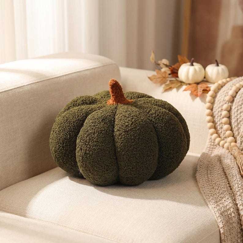 May include: A round, olive green pumpkin-shaped pillow made of textured, fuzzy sherpa fabric. The pillow features deep, vertical segments and a small, upright stem in a contrasting burnt orange color, creating a soft, plush, and rounded appearance.