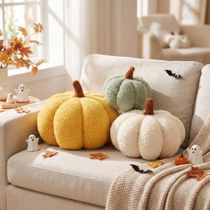 May include: Three plush pumpkin-shaped pillows in yellow, sage green, and cream, each featuring a brown stem. The pillows are arranged on a light-colored sofa alongside small white ghost figurines, scattered autumn leaves, and black bat cutouts.