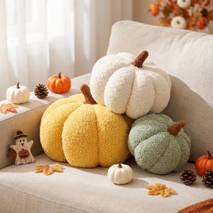 May include: Three plush pumpkin-shaped pillows in white, yellow, and sage green with textured sherpa-like fabric and brown stems. The pillows are arranged on a sofa alongside small decorative pumpkins, pinecones, and a ghost-shaped ornament.