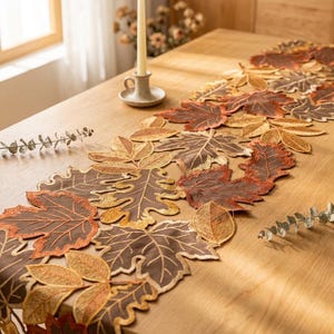 Pode incluir: Um caminho de mesa com um arranjo intrincado de folhas de outono bordadas em tons de castanho, laranja e dourado. O design consiste em várias formas de folhas com costuras detalhadas, criando uma aparência texturizada e em camadas.