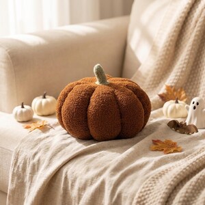 May include: A large, round, brown, textured pumpkin-shaped pillow with a light green stem. The pillow is surrounded by two small white decorative pumpkins, a small white ghost figurine, a small wooden bat, and several scattered autumn leaves.