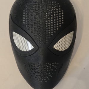 May include: A black full-face mask with a textured surface featuring a pattern of small circular perforations across the forehead and chin areas. The mask includes two almond-shaped eye openings covered with a white mesh material.