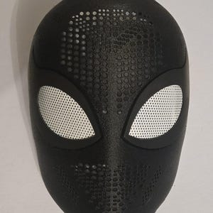 Könnte beinhalten: Eine schwarze Vollgesichtsmaske mit einer texturierten Oberfläche und einem perforierten Muster im Stirn- und Kinnbereich. Die Maske verfügt über zwei mandelförmige Augenöffnungen, die mit weißem Netzstoff bedeckt und schwarz umrandet sind.