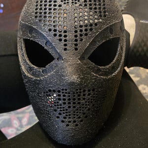 May include: A black, textured, full-face mask with a perforated pattern covering the forehead and mouth areas. The mask features two large, almond-shaped eye openings and a small, distinct red and white accent mark near the lower left side.