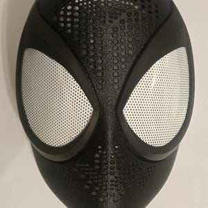 Könnte beinhalten: Eine schwarze Vollmaske mit strukturierter Oberfläche und zahlreichen kleinen kreisförmigen Perforationen im Stirn- und Kinnbereich. Die Maske verfügt über zwei große, mandelförmige Augenöffnungen, die mit einem feinen, silberfarbenen Netz bedeckt sind.