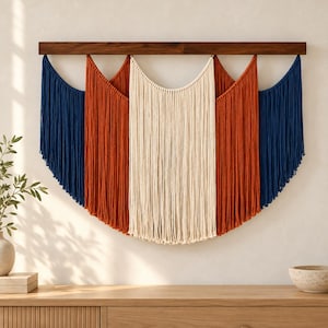 May include: A wall hanging featuring long, vertical cotton cords suspended from a horizontal dark wood bar. The design consists of five distinct sections of color arranged in a symmetrical pattern: navy blue, burnt orange, cream, burnt orange, and navy blue.