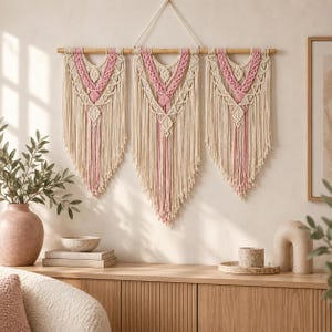 May include: A wall hanging featuring three macrame panels suspended from a wooden rod. The design uses cream and dusty pink cotton cord in intricate knotted patterns, with long fringe detailing at the bottom of each section and a hanging cord.