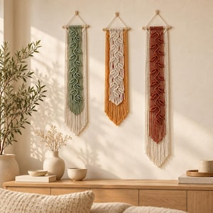 Peut inclure: Trois décorations murales en macramé suspendues à des tiges en bois. Chaque pièce présente un motif central de nœuds en forme de feuilles en vert sauge, jaune moutarde ou terre cuite, encadré par des franges et des cordons crème.