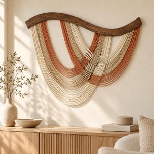 Puede incluir: Un tapiz de pared con una barra superior de madera oscura curvada y múltiples hilos drapeados de color crema y terracota. Los cordones están dispuestos en bucles superpuestos y estratificados que crean un patrón fluido y simétrico.