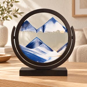 May include: A circular black frame holds a glass disc containing layers of blue, white, and gold sand. The sand is arranged in mountain-like peaks within the liquid-filled chamber, supported by a rectangular black base that allows for rotation.