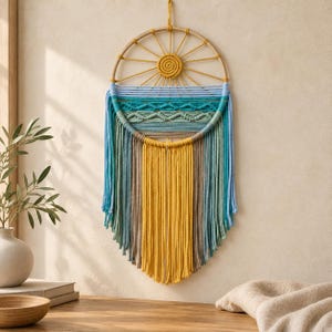 May include: A circular macrame wall hanging featuring a central sun motif made of yellow cord. The design incorporates horizontal rows of knotted patterns in shades of blue, green, and tan, with long, flowing fringe tassels hanging from the bottom.