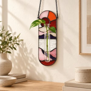 May include: A vertical oval stained glass wall hanging featuring a sunset-inspired pattern in shades of orange, pink, purple, and white. A clear glass test tube is mounted in the centre, holding a green plant cutting with two leaves.