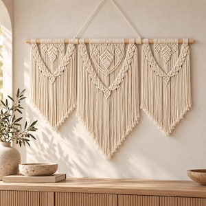 May include: A large off-white macrame wall hanging suspended from a horizontal wooden dowel. The piece features three distinct sections with intricate knotted patterns, layered geometric shapes, and long, flowing fringe that tapers to points.