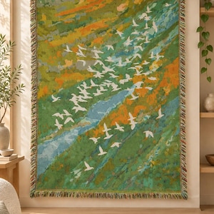 May include: A rectangular woven throw blanket featuring a landscape scene with a flock of white birds flying over a blue river and green and orange hills. The blanket has a fringed edge with multicoloured threads and is hung on a wooden rod.
