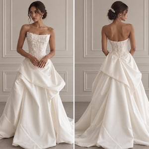 Peut inclure: Une robe de mariée bustier ivoire dotée d'un corsage style corset avec des broderies florales et des perles complexes. La jupe est confectionnée en satin lisse avec des panneaux drapés superposés créant une silhouette volumineuse et longue.