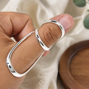 May include: A silver-colored metal splint ring featuring a smooth, polished finish. The design consists of two circular bands connected by a long, curved bar that extends along the side of the finger, providing a sleek and continuous appearance.