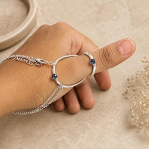 May include: A silver hand chain featuring a circular ring component connected to a smaller ring by a thin silver chain. Both rings are set with a small, round, dark blue gemstone. The hand chain includes a delicate silver link chain with a lobster clasp.