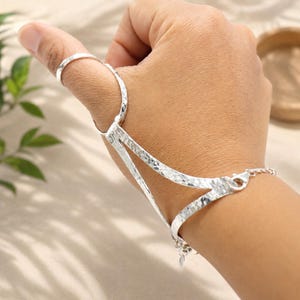 May include: A silver hand chain featuring a hammered texture. The piece consists of a ring that encircles the thumb, connected by a V-shaped metal structure to a bracelet that fastens around the wrist with a small lobster claw clasp.