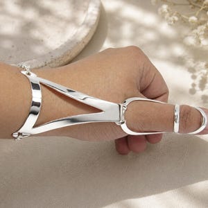 May include: A polished silver-tone hand harness featuring a wide, curved wrist cuff connected by a V-shaped metal bridge to a ring that encircles the thumb. The piece is constructed from smooth, reflective metal with a continuous, sleek design.