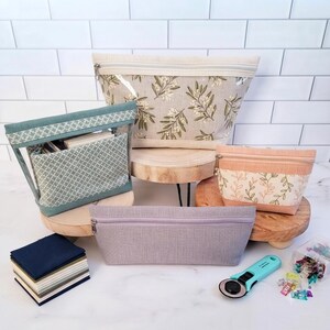 Può includere: Una collezione di quattro pochette in tessuto con cerniera di varie dimensioni e fantasie, tra cui una grande stampa con rami d'ulivo, un motivo geometrico verde acqua, una stampa con viticci color pesca e una pochette lavanda tinta unita, insieme a quadrati di tessuto e una taglierina rotante.