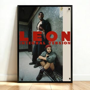 May include: A framed film poster featuring a man in a dark coat and sunglasses standing behind a seated person wearing a red beanie and a green jacket. The text LEON INTEGRAL VERSION is printed in large red letters across the centre of the image.