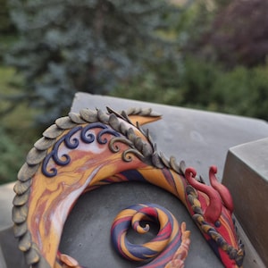 Könnte beinhalten: Eine zusammengerollte Drachenfigur mit einem marmorierten Muster in Orange, Gelb, Violett und Rot. Das Wesen hat strukturierte, schuppenartige Kämme auf dem Rücken, dunkelviolette Wirbelakzente und glatte rote Hörner am Kopf.